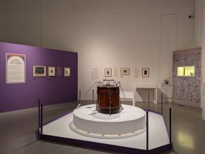 A round wooden tub with metal rods and cords projecting from its lid, displayed on a white plinth in a museum gallery with framed pictures on purple and white walls