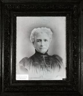 Black-and-white portrait of an older woman with white hair and a high-necked dark dress, in an ornate carved wooden frame