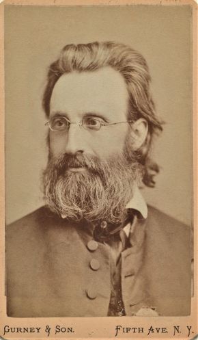 Sepia studio portrait of a bearded man with long hair and wire-rimmed glasses