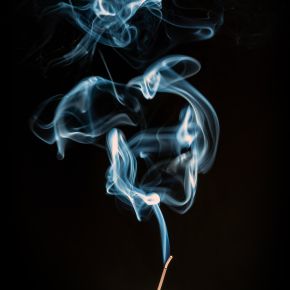 Curling blue-white wisps of smoke rising from the glowing tip of an incense stick against a black background