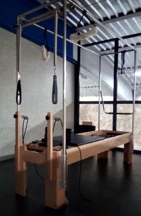 A wooden Pilates bed with a padded sliding carriage, under a metal frame hung with straps and springs, in an empty studio