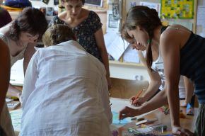 Several people standing around a table, drawing together on a large sheet of paper with coloured pencils and pens