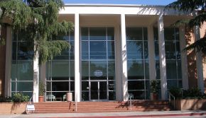Glass-fronted entrance of a low building with white columns, trees and brick steps