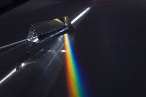 A clear triangular glass prism on a dark surface, with a band of rainbow colours spreading out beside it