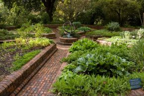 Raised brick garden beds of leafy herbs beside a curving brick path, with a sundial in the background