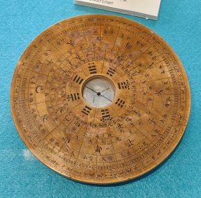 A round wooden compass plate with a small magnetic needle at its centre, surrounded by rings of Chinese characters and trigrams