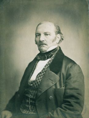Black-and-white photographic portrait of a balding man with a moustache, wearing a dark coat and checked waistcoat