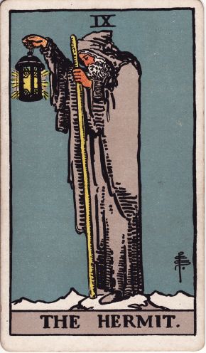 A tarot card showing a grey-bearded hermit in a long cloak, holding up a lantern and a staff, with the title The Hermit