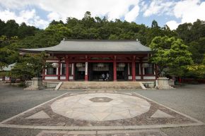 A red and white wooden temple hall with a grey tiled roof, reached by stone steps across a paved courtyard, with forested hills behind