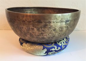 A bronze-coloured metal bowl engraved with script, resting on a blue and gold cushion
