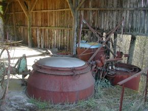 A large rust-red metal still with pipes and tanks, standing in an open timber shed