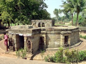 A low, circular stone temple with carved figures set into niches in its outer wall, among trees and palms