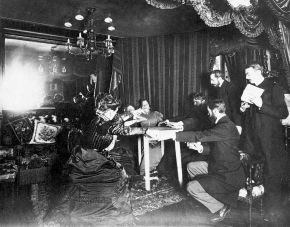 Black-and-white photograph of a group of people in late nineteenth-century dress gathered around a small table in a curtained parlour