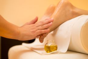 Close-up of a practitioner's hands working on the sole of a bare foot resting on a towel, with a small glass jar of oil beside it